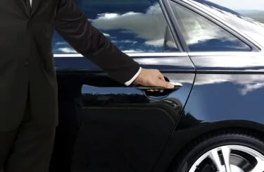 Jet Charter A person dressed in a black suit is opening the door of a sleek, black car. The car's exterior reflects the blue sky and clouds, hinting at the precious cargo inside. Only the person's arm and part of their body are visible. Jetvia