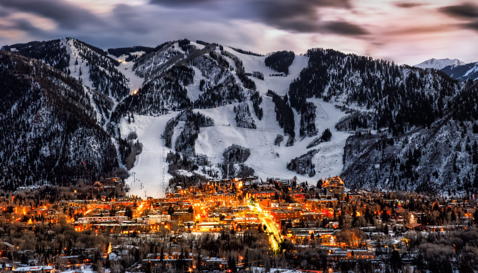 Jet Charter A twilight view of a snow-covered mountain town with glowing lights amidst the peaks, highlighting a ski resort and densely packed buildings, accessible by private jet charter. Jetvia