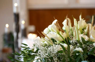 Jet Charter A close-up of a floral arrangement featuring white calla lilies, baby's breath, and various greenery. Blurred candles and a softly lit interior create a serene, elegant atmosphere in the background, subtly reminiscent of memorials honoring human remains. Jetvia