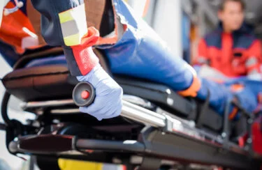 Jet Charter A close-up shot shows a gloved hand gripping a handle on a stretcher. In the background, a person in red and blue emergency medical attire is slightly blurred, indicating they are in motion, likely inside an air ambulance. Jetvia