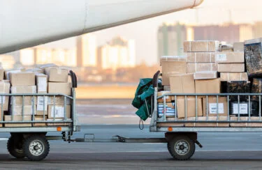Jet Charter Two baggage carts loaded with numerous cargo packages and boxes are parked on the tarmac next to a charter airplane. The background features a cityscape with tall buildings. Jetvia