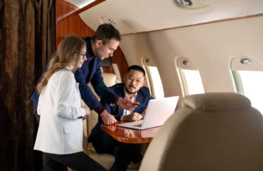Jet Charter Three professionals are engaged in a discussion while looking at a laptop screen inside a Group Air Charter jet. One man is pointing at the screen while the other man and a woman listen attentively. They appear to be working collaboratively during the flight. Jetvia