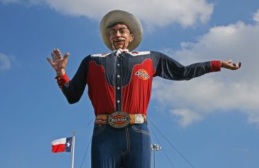 Jet Charter Dallas A large, animated fiberglass statue of a cowboy with a giant hat, welcoming smile, and outstretched arms under a blue sky with clouds, symbolizing Texan culture. A Texas flag waves in the background. Nearby, a private jet charter is ready for departure. Jetvia