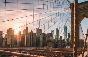 Jet Charter Sunset view of New York City skyline through the cables and structure of the Brooklyn Bridge, highlighting the architectural details and urban scenery. Consider enhancing your experience with a Private Jet Charter. Jetvia