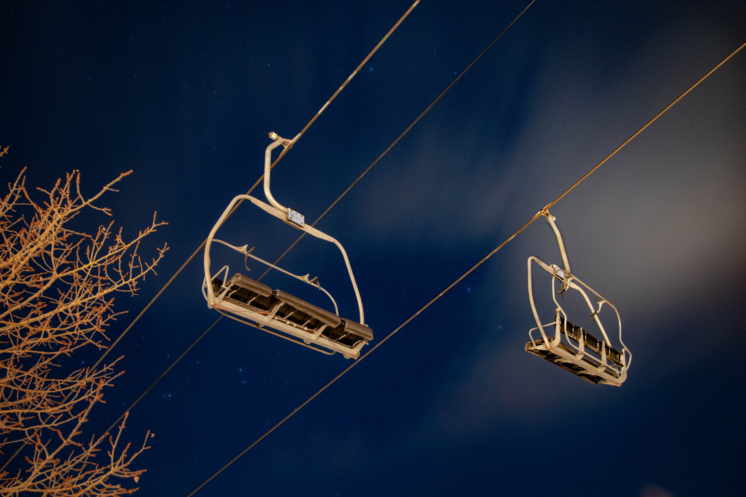 Jet Charter Two empty ski lift chairs suspended against a night sky, with tree branches on the left and a glowing moon partially obscured by clouds. Jetvia