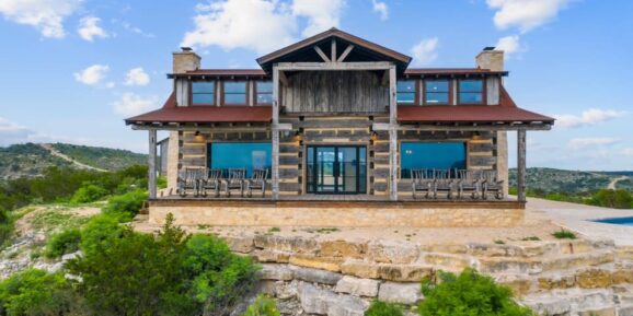 Jet Charter A large, rustic log cabin with a stone foundation sits atop a rocky cliff on the picturesque Ox Ranch. Surrounded by greenery, the cabin boasts large windows, a metal roof, and a wraparound porch with rocking chairs, offering views of the hilly landscape under a partly cloudy sky. Jetvia