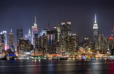 Jet Charter Private Jet Charter: Nighttime view of New York City skyline illuminated against a dark sky, featuring the Empire State Building and multiple skyscrapers with reflections on the water. Jetvia