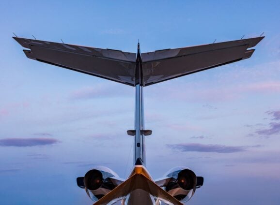 Jet Charter A rear shot of an airplane with the horizontal and vertical stabilizers visible, set against a backdrop of a clear sky with soft, pinkish clouds during either sunrise or sunset. This description captures the serene beauty and tranquil moment experienced at high altitudes. Jetvia