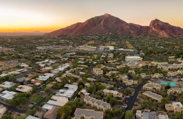 Jet Charter Scottsdale Aerial view of a sprawling suburban neighborhood in Scottsdale with a backdrop of large mountains at sunset. The landscape includes numerous houses, green spaces, and winding roads, with the mountains bathed in a warm, golden light from the setting sun. Jetvia