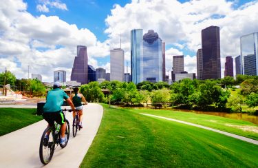 Jet Charter Houston Two people ride bicycles on a path through a green park in Houston, Texas, with the skyline in the background on a sunny day with white clouds. The park is lush and vibrant, and the towering skyscrapers create a striking contrast against the natural scenery. Jetvia