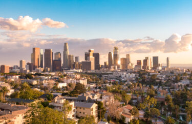 Jet Charter A panoramic view of the Los Angeles skyline at sunset. The city's downtown buildings rise prominently against a backdrop of a partly cloudy sky, with the setting sun casting a warm glow. In the foreground, Los Angeles's residential neighborhoods nestle amidst lush green trees. Jetvia