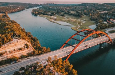 Jet Charter Austin Aerial view of a red arched bridge spanning a wide river with a golf course on one side and rocky cliffs on the other. The skyline of Austin is faintly visible in the far background. Vehicles can be seen crossing the bridge, and the scene is bathed in warm light. Jetvia