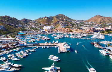 Jet Charter Aerial view of a bustling marina with numerous docks and boats in a vibrant blue body of water. Surrounding the marina are buildings, including hotels and residences, nestled among rugged, green hills under a clear, blue sky—a picturesque sight reminiscent of flying Houston to Cabo San Lucas. Jetvia