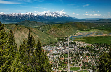 Jet Charter A scenic view of Jackson Hole showcases a picturesque mountainous landscape with a small town nestled in the valley. Snow-capped mountains are visible in the background under a blue sky. Lush green hills and pine trees surround the town below, perfect for travel and tourism. A river runs through the landscape to the right. Jetvia