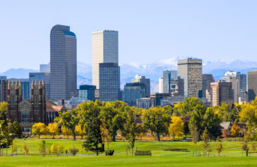 Jet Charter Skyline of Denver with modern skyscrapers and historic buildings surrounded by greenery, trees in fall colors, and a clear blue sky. Snow-capped mountains are visible in the background. Jetvia