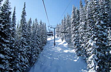 Jet Charter A snow-covered mountain slope with dense pine trees, seen from a ski lift ascending toward the top. An eagle soars gracefully above under the clear blue sky, while the ski lift cables stretch into the distance. Jetvia