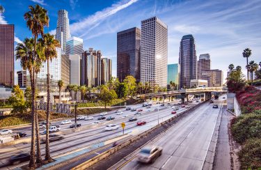 Jet Charter A bustling freeway with multiple lanes of vehicles stretches through downtown Los Angeles, bordered by tall skyscrapers and palm trees under a bright, clear blue sky. Trails of clouds streak across the sky, enhancing the vibrant urban travel scene. Jetvia