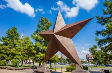 Jet Charter Austin A large bronze star sculpture adorns a park setting, framed by trees and a clear blue sky with scattered clouds. To the left stands a flagpole, while buildings and parked cars are visible in the distance, reminiscent of similar scenes from Atlanta to Austin. Jetvia