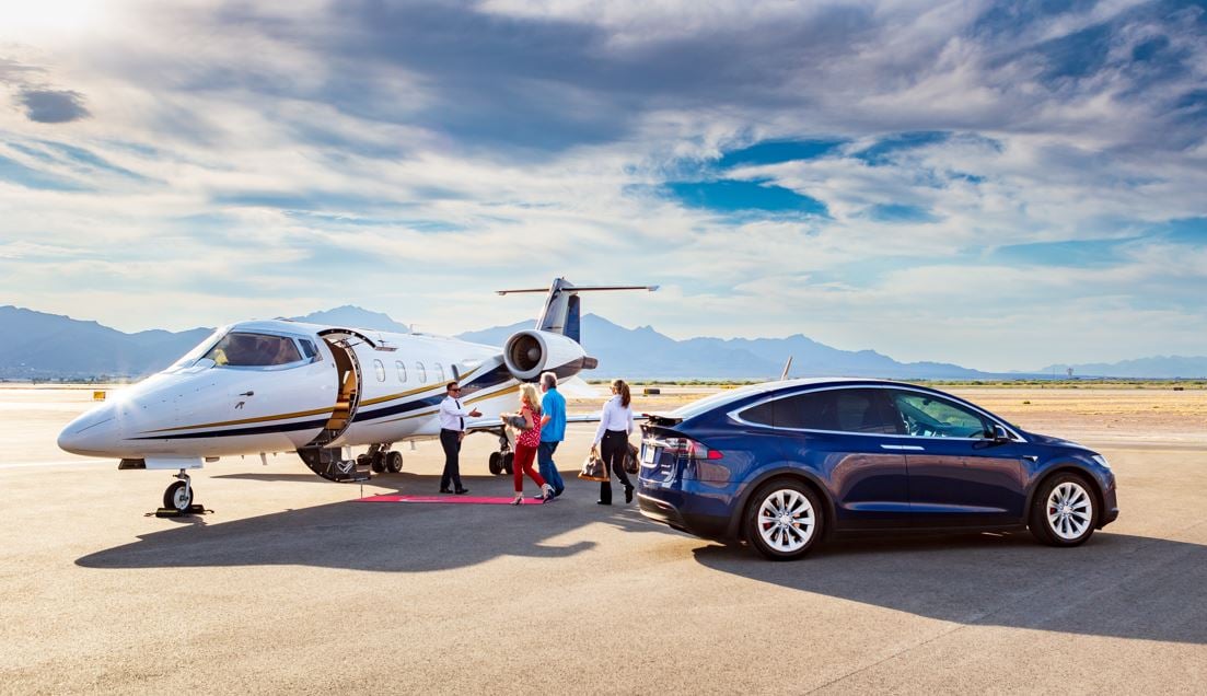 Jet Charter A private jet is parked on an airstrip with mountains in the background, ready for its next journey. A group of people are boarding the jet via a staircase. Nearby, a blue Tesla SUV sits on the tarmac under the bright sky. This could be an ideal scenario for travelers looking for Empty Legs deals. Jetvia
