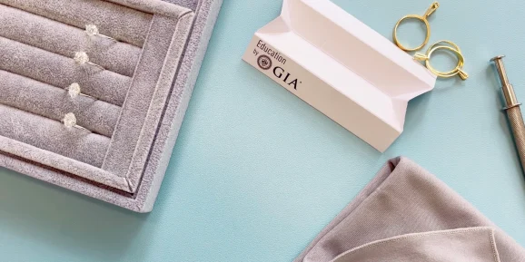Jet Charter A close-up of a custom jewelry workspace featuring a gray ring holder with diamond rings, a GIA booklet titled "Education by GIA," a pair of gold magnifying loops, a gray jewelry cloth, and a metal tool against a light blue background. Jetvia