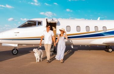 Jet Charter A man and woman walk hand-in-hand on a tarmac with a white dog beside them. They are exiting their private jet under a bright blue sky. The man is wearing a white shirt and beige pants, while the woman wears a white dress and a sun hat, epitomizing luxurious air travel. Jetvia