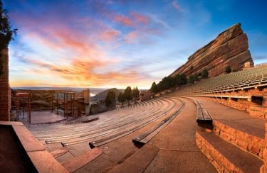 Jet Charter Denver A panoramic view of the Red Rocks Amphitheatre at sunset just outside Denver. The empty seating area curves around a stage positioned amidst towering red sandstone formations. The sky above is vivid with shades of orange, pink, and blue as the sun sets in the distance. Jetvia