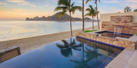 Jet Charter Infinity pool overlooking a serene beach with calm waters and a mountainous horizon at sunset. Palm trees line the edge of the pool, and a stone wall fountain adds a touch of elegance to the tranquil scene. Experience ultimate relaxation in one of our luxury villas in Cabo. Jetvia