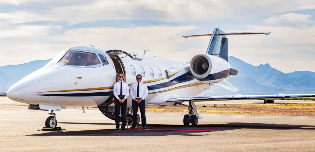 Jet Charter Two pilots, one female and one male, stand confidently in front of a private jet charter on a sunny tarmac with mountains in the background. Jetvia
