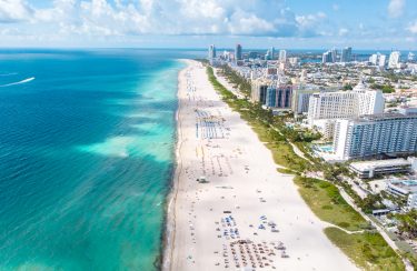 Jet Charter Miami Aerial view of a lengthy beach with white sand and turquoise waters, lined with colorful beach umbrellas and people relaxing. High-rise buildings and a cityscape, reminiscent of the journey from Dallas to Miami, are visible in the background under a partly cloudy sky. Jetvia