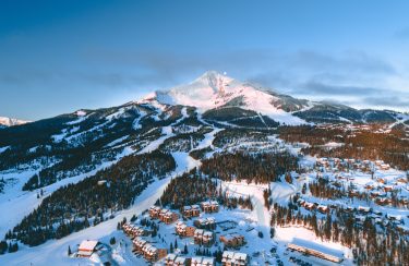 Jet Charter Big Sky A snow-covered mountain with ski slopes and trails is surrounded by forested areas under a vast Big Sky. At the mountain's base, clusters of wooden chalets and lodges nestle cozily. The sky is clear with a hint of clouds at the peak, creating a serene and picturesque scene. Jetvia