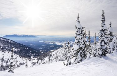 Jet Charter Big Sky A scenic view of a snowy mountain landscape under the bright Big Sky. Tall pine trees covered in snow are in the foreground, while distant mountains and valleys stretch out below. The sun shines softly, creating a serene and tranquil atmosphere. Jetvia