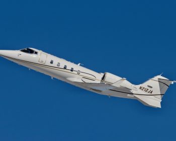 Jet Charter A white private jet, likely a Learjet 60, with registration number N212JA is captured flying at an angle against a clear blue sky. The aircraft embodies the pinnacle of aviation, featuring a sleek design with gold and black detailing along its fuselage. Its landing gear is retracted, indicating it is in flight. Jetvia