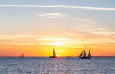 Jet Charter Three sailboats glide on the calm ocean under a vibrant Key West sunset sky. The sun, partially set on the horizon, casts a golden glow over the water, blending into hues of orange, pink, and blue. The distant horizon blurs seamlessly with the sky above. Jetvia