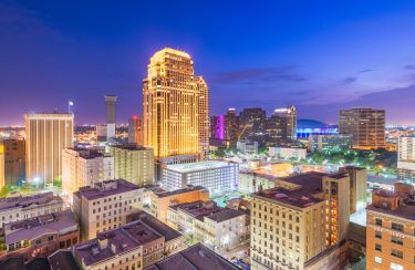 Jet Charter A nighttime aerial view of the New Orleans skyline with illuminated skyscrapers and buildings. One prominent building is brightly lit, centrally dominating the scene. The sky has a gradient from blue to purple, and city lights twinkle, highlighting the urban landscape. Jetvia
