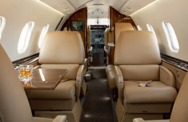Jet Charter The interior of a private jet, perfect for exploring exclusive routes, features luxurious beige leather seats and a wooden table with two glasses. The cockpit is visible in the background, flanked by curved walls and windows that allow natural light to flood in. Jetvia