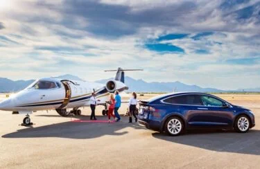 Jet Charter A private jet rests on the airport runway, with mountains majestically framing the scene. As a group begins boarding, a blue electric SUV—prize from the recent Greens Sweepstakes—waits nearby. The sky above is partly cloudy, adding to the picturesque moment. Jetvia