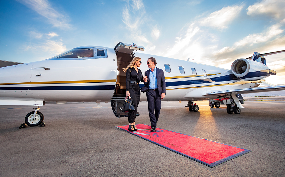 Jet Charter A well-dressed man and woman exit a Bombardier Learjet 60 onto a red carpet at sunset, holding hands and smiling. The scene reflects luxury and exclusivity, with the jet and sky in the background. Jetvia