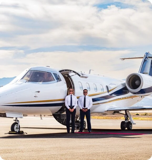 Jet Charter Two pilots in uniform stand on a red carpet in front of a white private jet parked on an airport runway under a partly cloudy sky. Jetvia