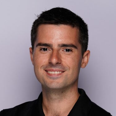 Jet Charter A man with short dark hair smiles with arms crossed, wearing a black polo shirt with a small "DETWA" logo, representing our team of experts against a plain light grey background. Jetvia