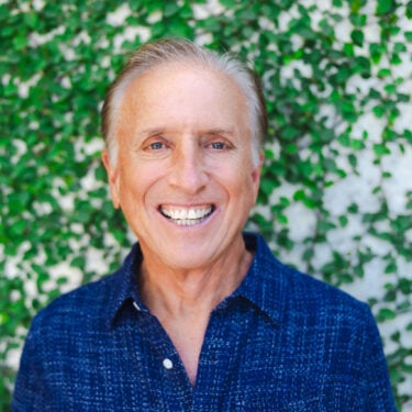 Smiling man in a blue shirt standing in front of a leafy green background.