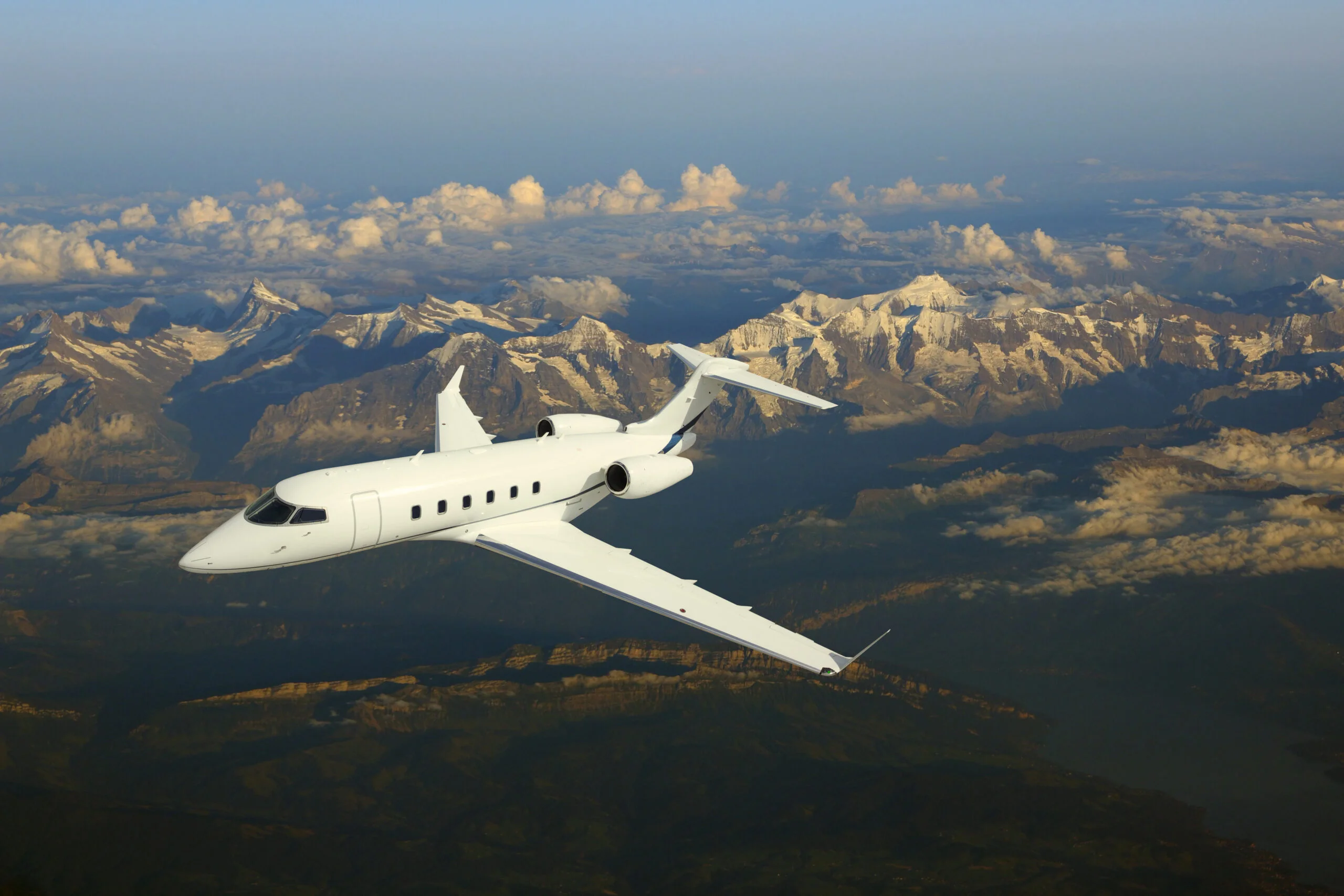 Jet Charter A white private jet from an experienced private jet charter company soars above snow-capped peaks, with scattered clouds and a clear sky providing a stunning mountain backdrop. Jetvia
