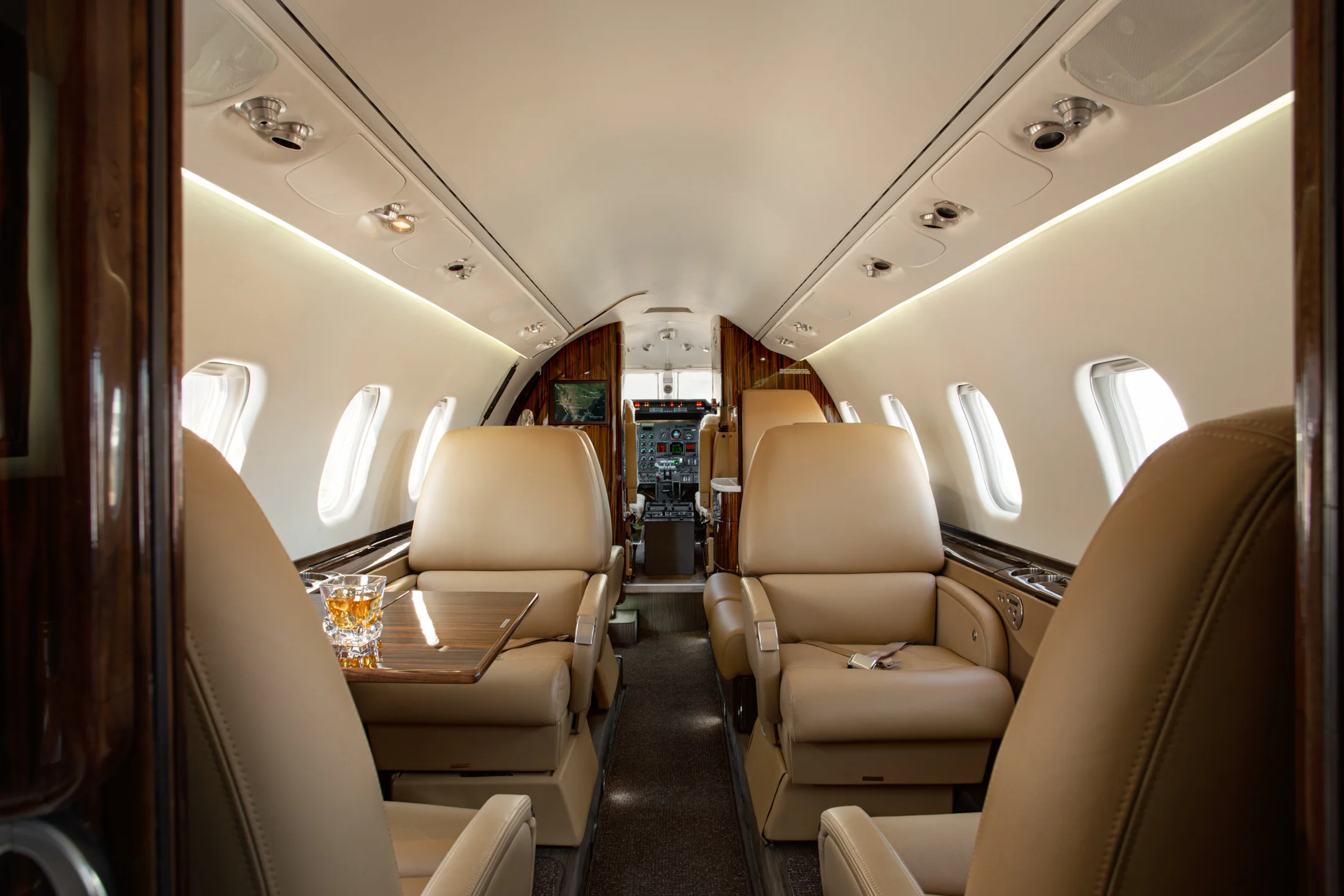 Jet Charter The interior of a private jet features plush beige leather seats, a small table with drinks, and large windows on both sides. The cockpit is visible at the front of the cabin. Jetvia