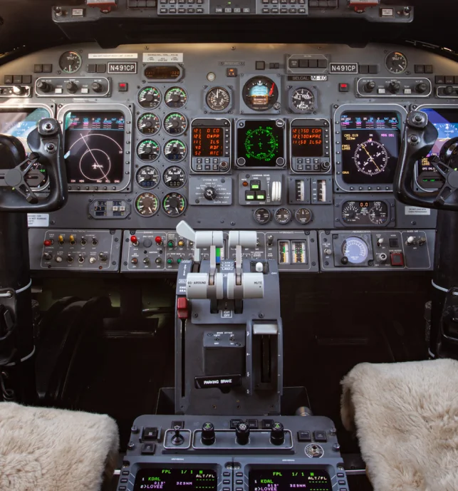 Jet Charter Cockpit of an aircraft featuring numerous instruments, gauges, buttons, and digital displays, with two sheepskin-covered pilot seats in the foreground. The controls and panels are detailed and well-lit. Jetvia