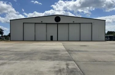 Jet Charter A large, closed white Jetvia maintenance facility sits on a wide, empty concrete apron under a partly cloudy blue sky. The hangar doors are shut, and there are no visible vehicles or people nearby. Jetvia