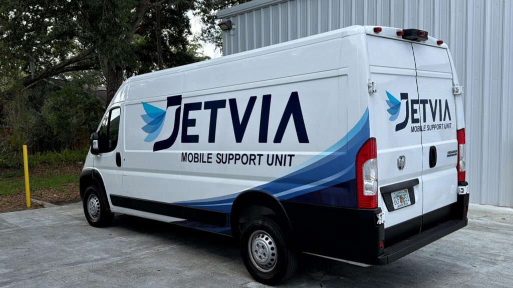 Jet Charter A white Jetvia van with "JETVIA MOBILE SUPPORT UNIT" and a blue wing logo is parked outside a metal maintenance facility, next to some trees. Jetvia