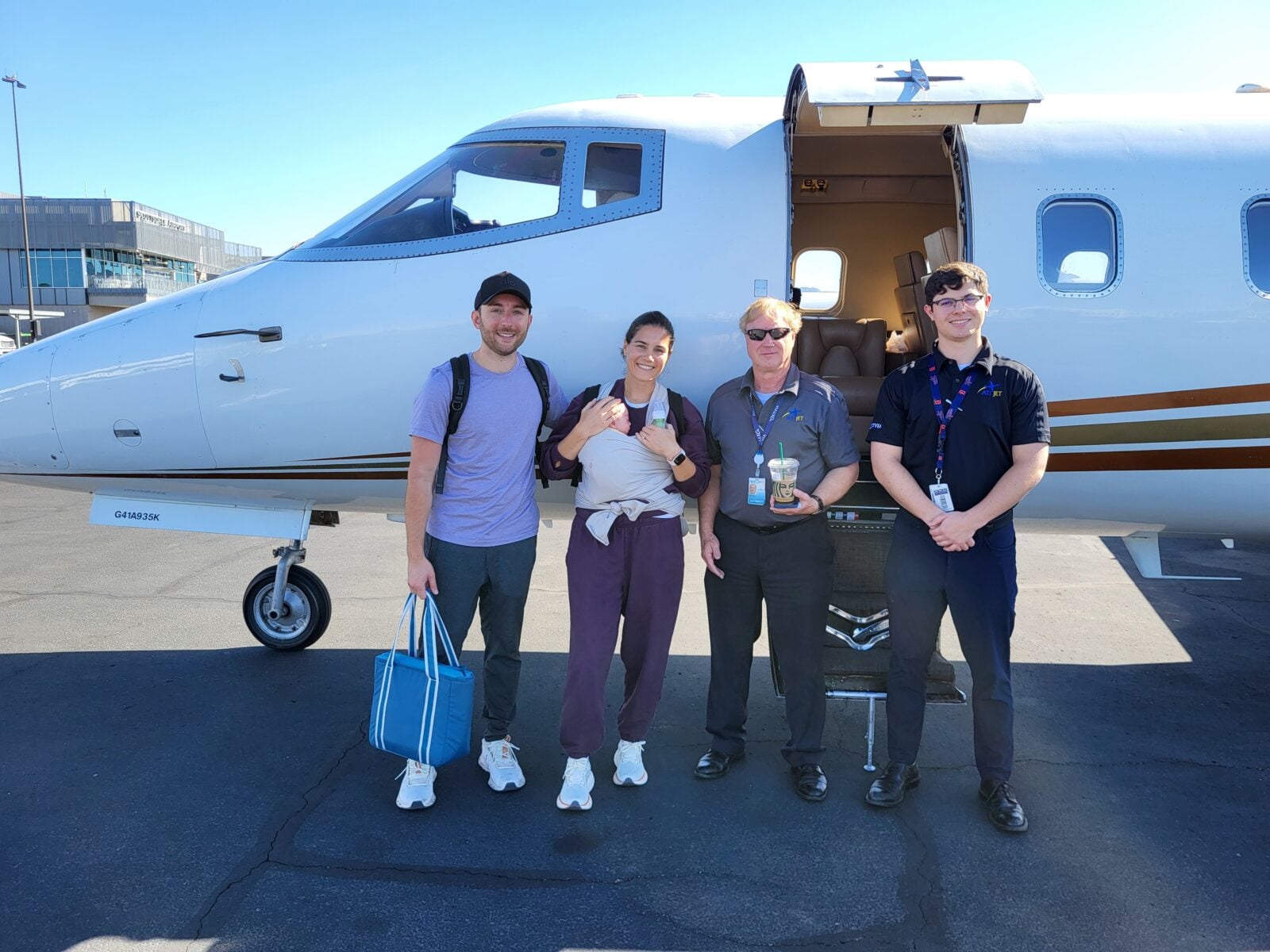 Jet Charter Four people stand smiling in front of a small private jet on an airport tarmac. Two, holding bags, are casually dressed; the other two wear uniforms. The jet’s door is open under a clear blue sky—an Elementor journey about to begin. Jetvia