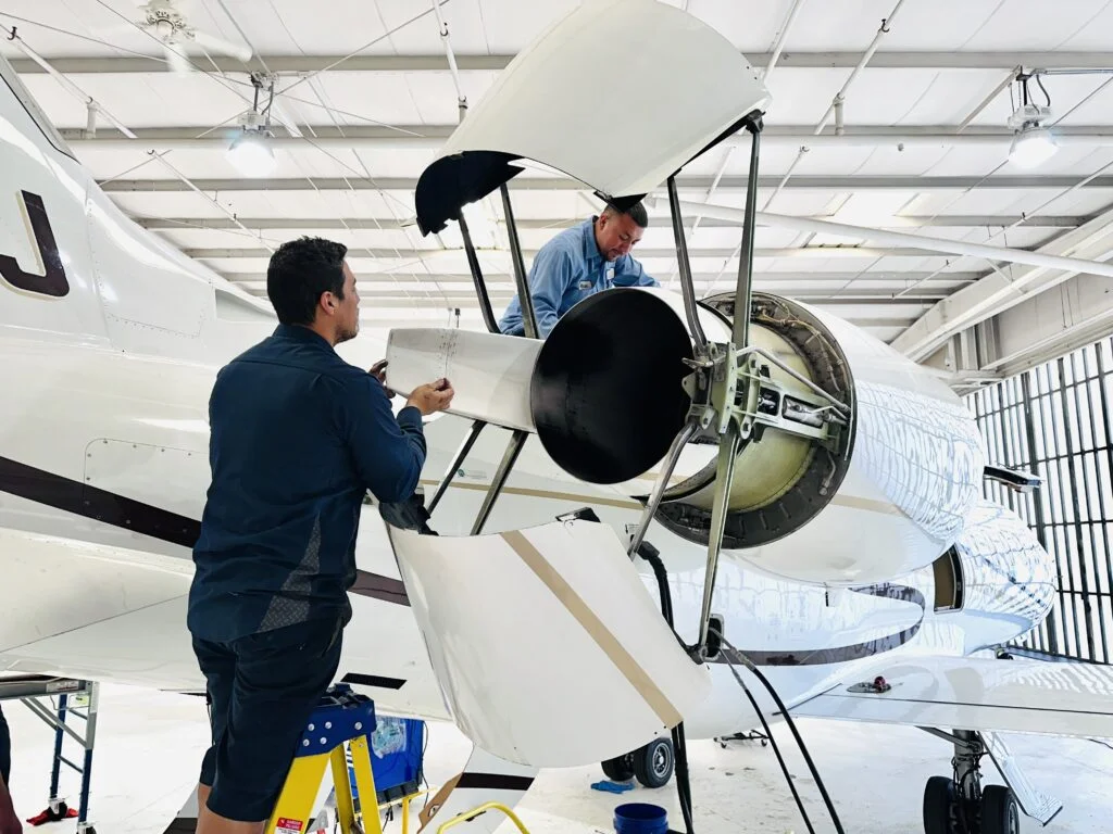 Jet Charter Two technicians from an experienced private jet charter company work on the exposed engine of a jet aircraft inside a well-lit hangar. One stands on a step stool while the other is inside the engine compartment, both focused on maintenance tasks. Jetvia