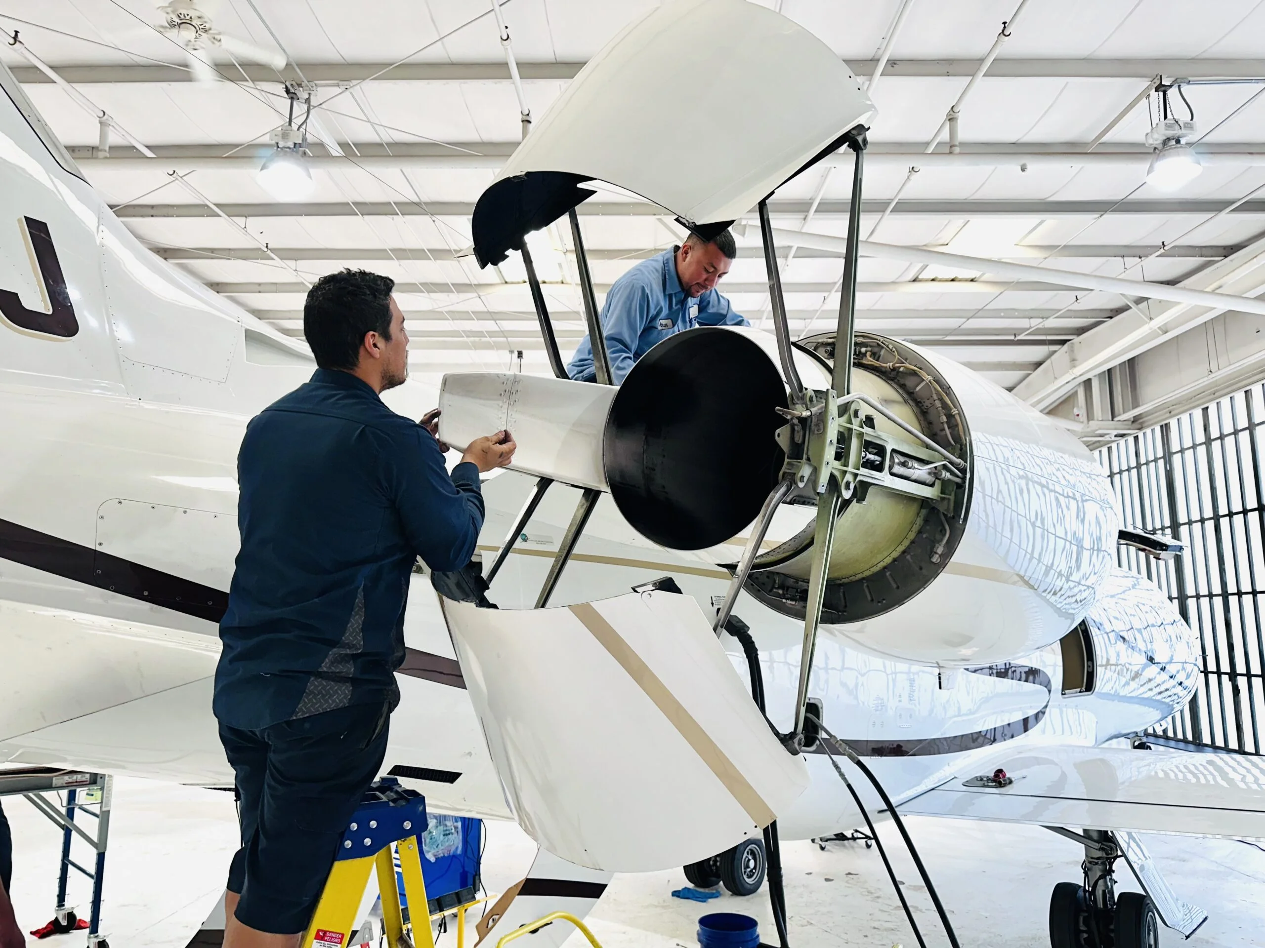 Jet Charter Two technicians from an experienced private jet charter company work on the exposed engine of a jet aircraft inside a well-lit hangar. One stands on a step stool while the other is inside the engine compartment, both focused on maintenance tasks. Jetvia