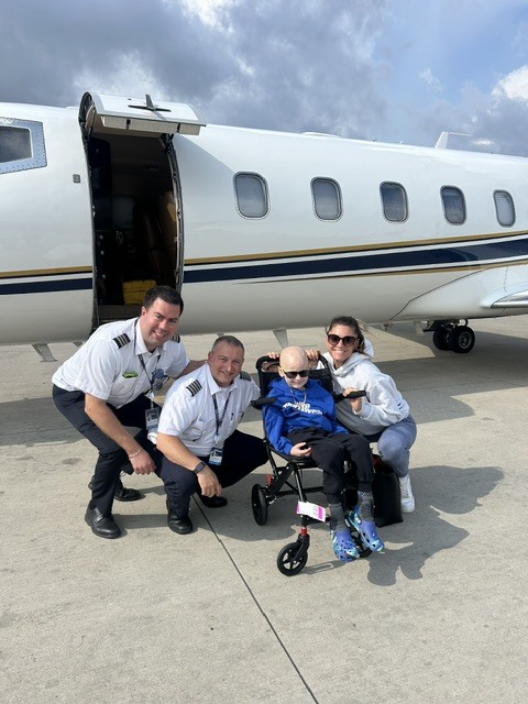 Jet Charter Two pilots and a woman kneel beside a smiling child in a wheelchair in front of a private jet on an airport tarmac under a partly cloudy sky. Jetvia