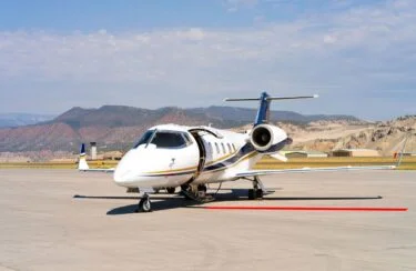 Jet Charter A small white private jet with an open door is parked on a sunny airport runway, ready for exclusive flights from New York to Los Angeles, with mountains and a partly cloudy sky in the background. Jetvia